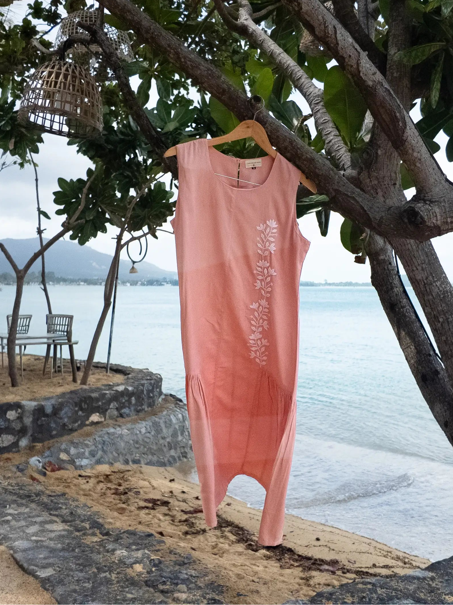 Pink dress hanging on a tree by the beach with chairs and tables in the background.
