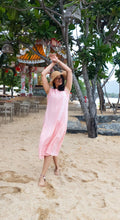 Woman in a pink dress standing on a sandy beach with trees and a scenic background.