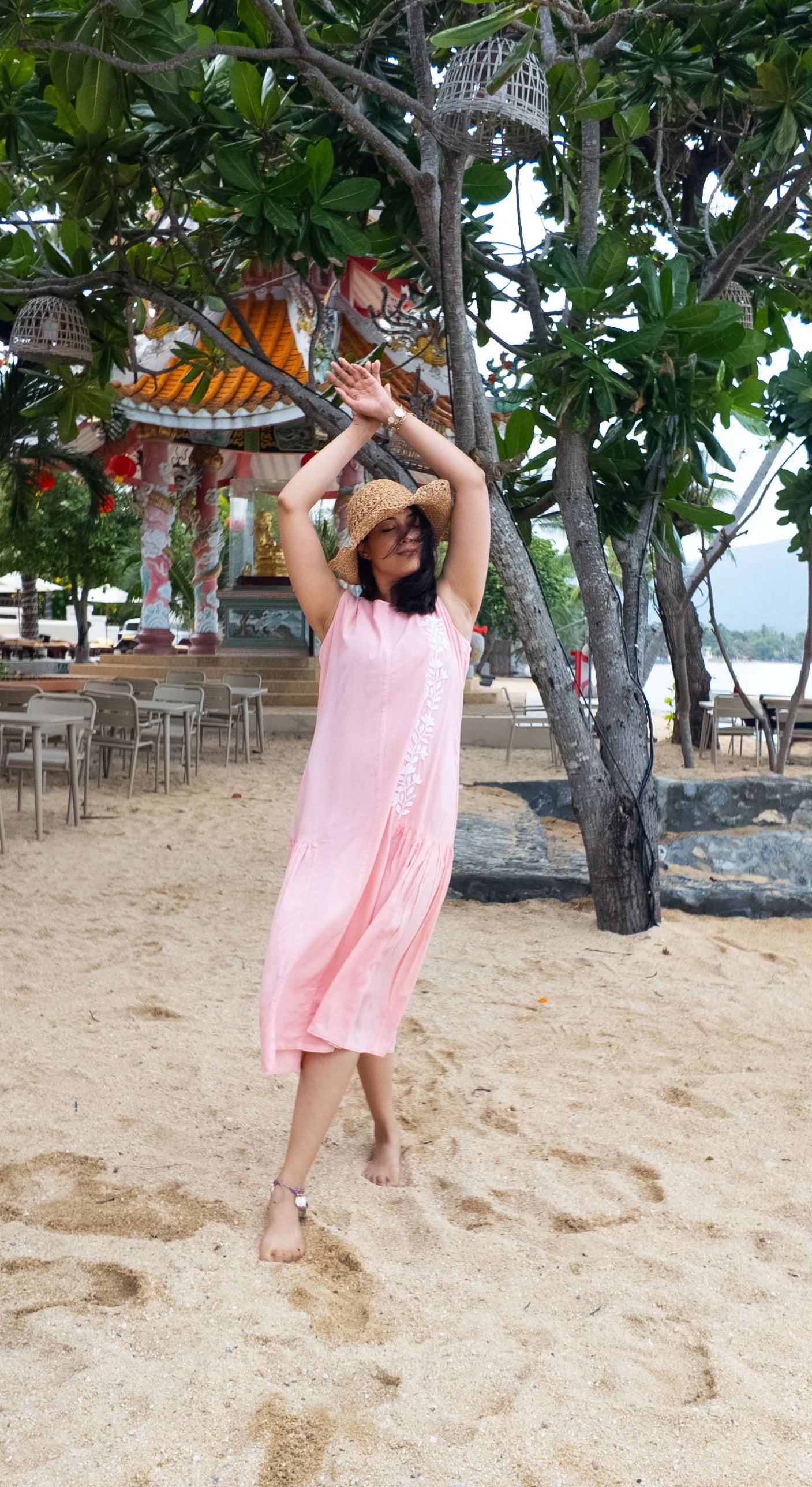 Woman in a pink dress standing on a sandy beach with trees and a scenic background.